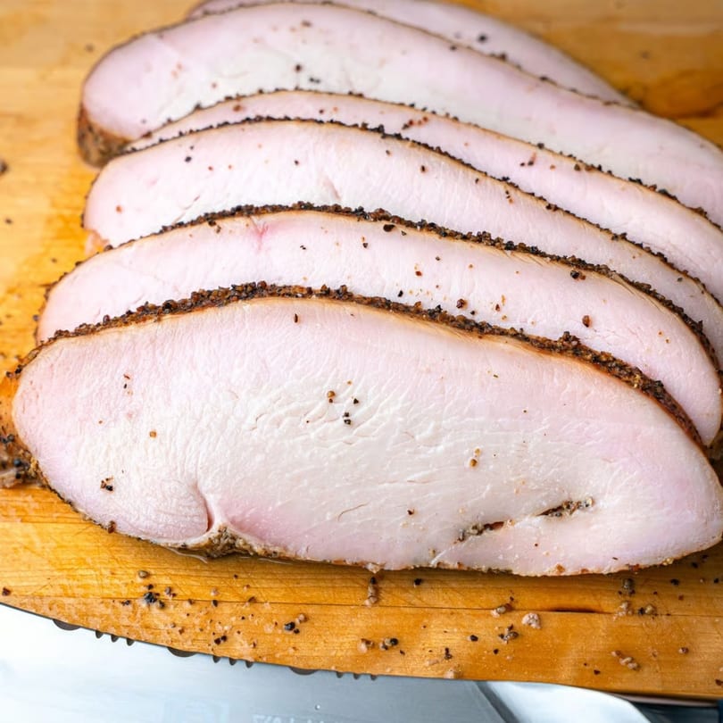 Thinly sliced, seasoned turkey breast on a wooden cutting board with a knife.